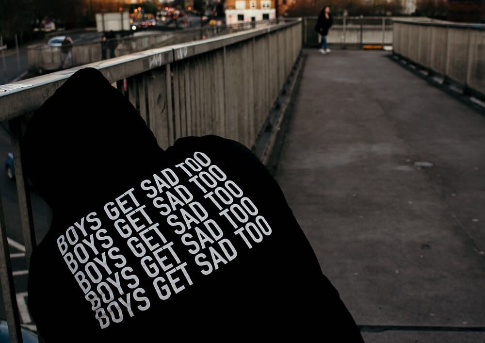 A man sitting on a bridge wearing a black hoodie with 'Boys get sad too' printed on the back.